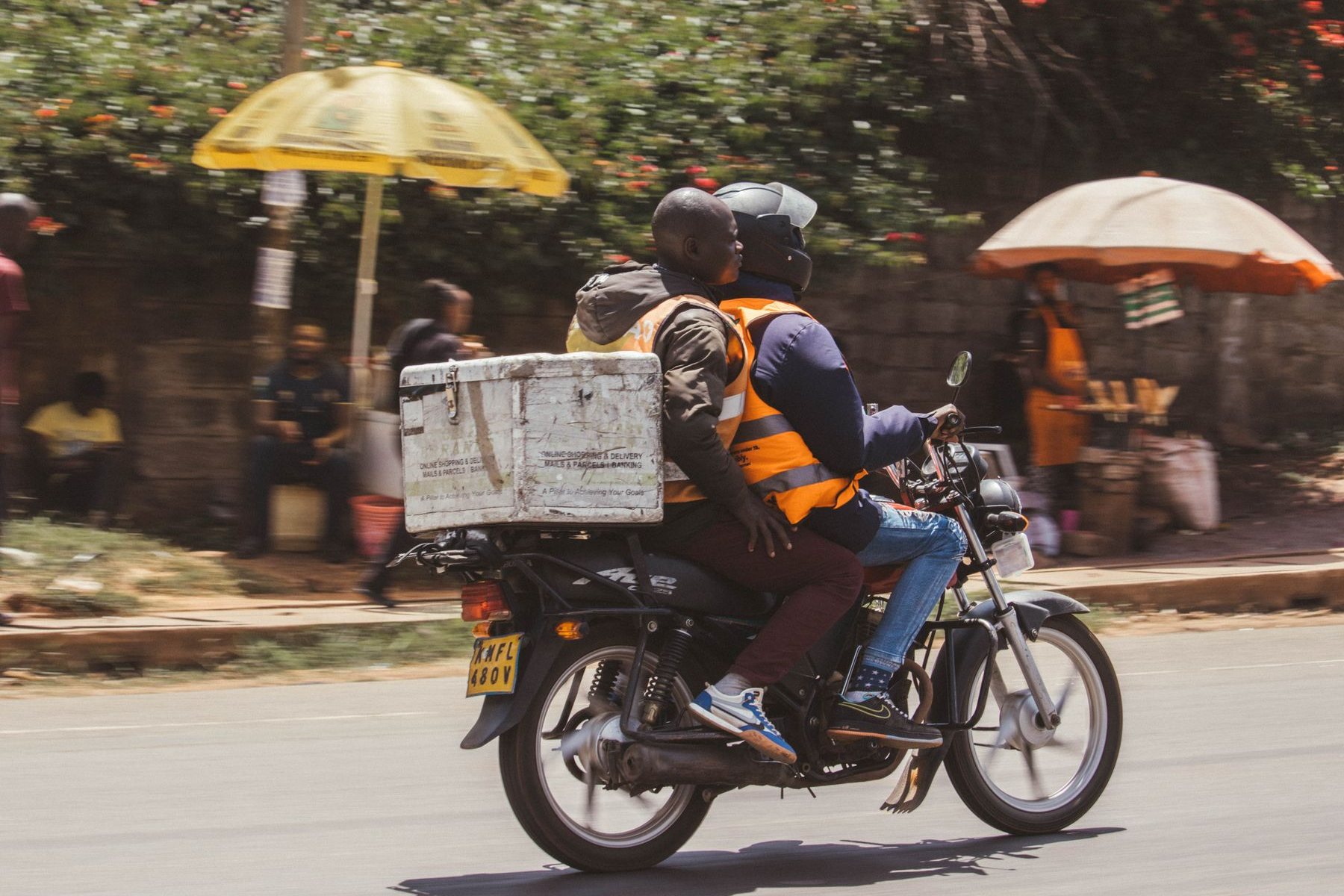 A Black Kenyan boda-boda courier in an orange hi-vis vest rides through a bright Nairobi street with a white delivery box strapped to the ba