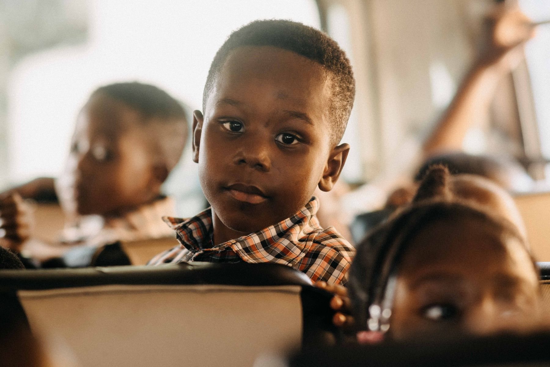A young boy in a checked shirt sits among other children riding a school bus in warm morning light