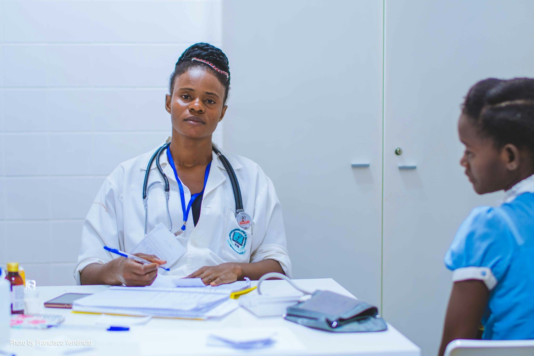 An African woman doctor in a white coat with stethoscope sits at her consultation desk writing notes while a young girl patient in a blue dr