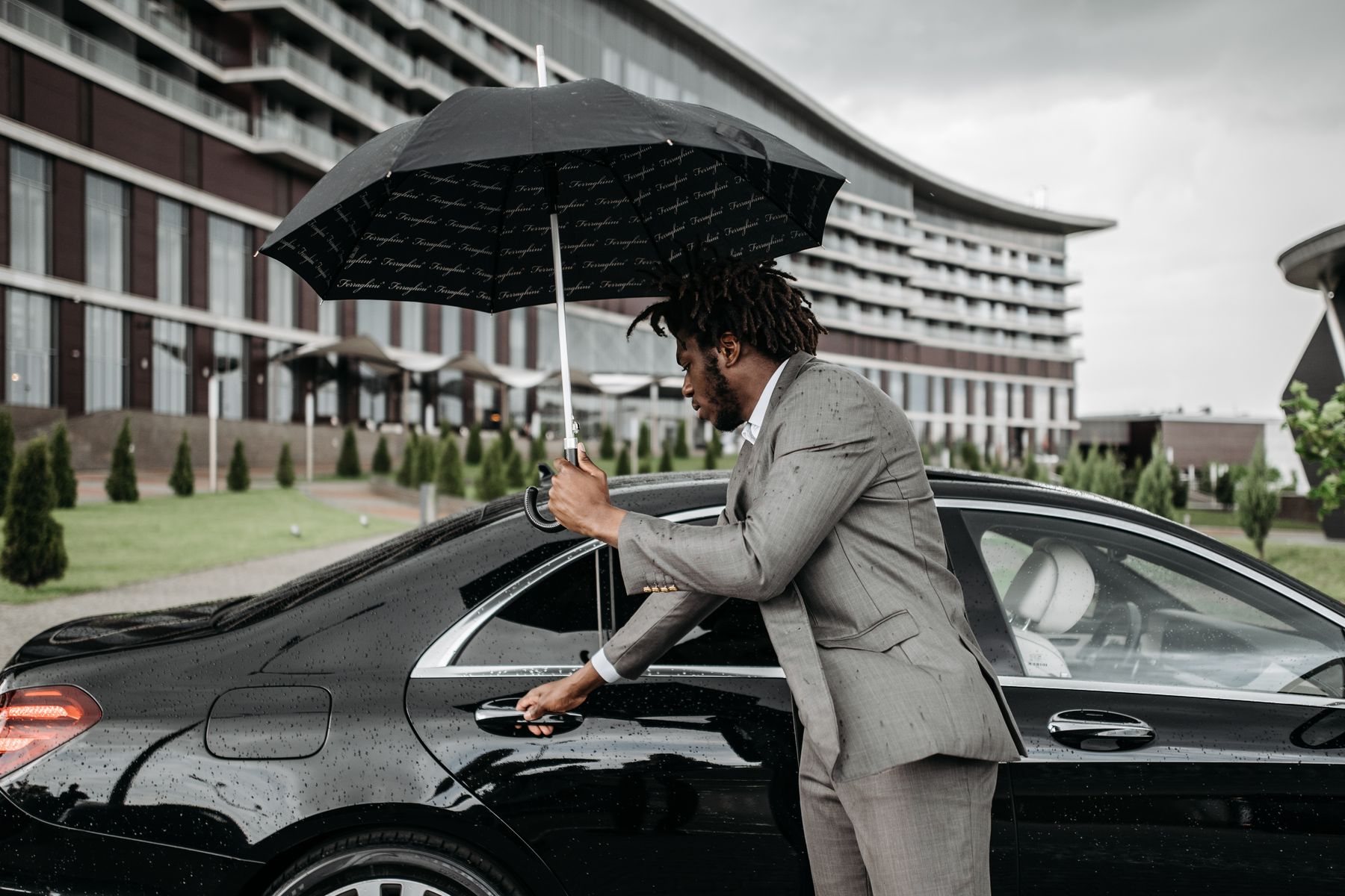 A Black concierge in a grey suit holds an umbrella and opens the door of a black sedan for a guest in front of a large modern hotel building