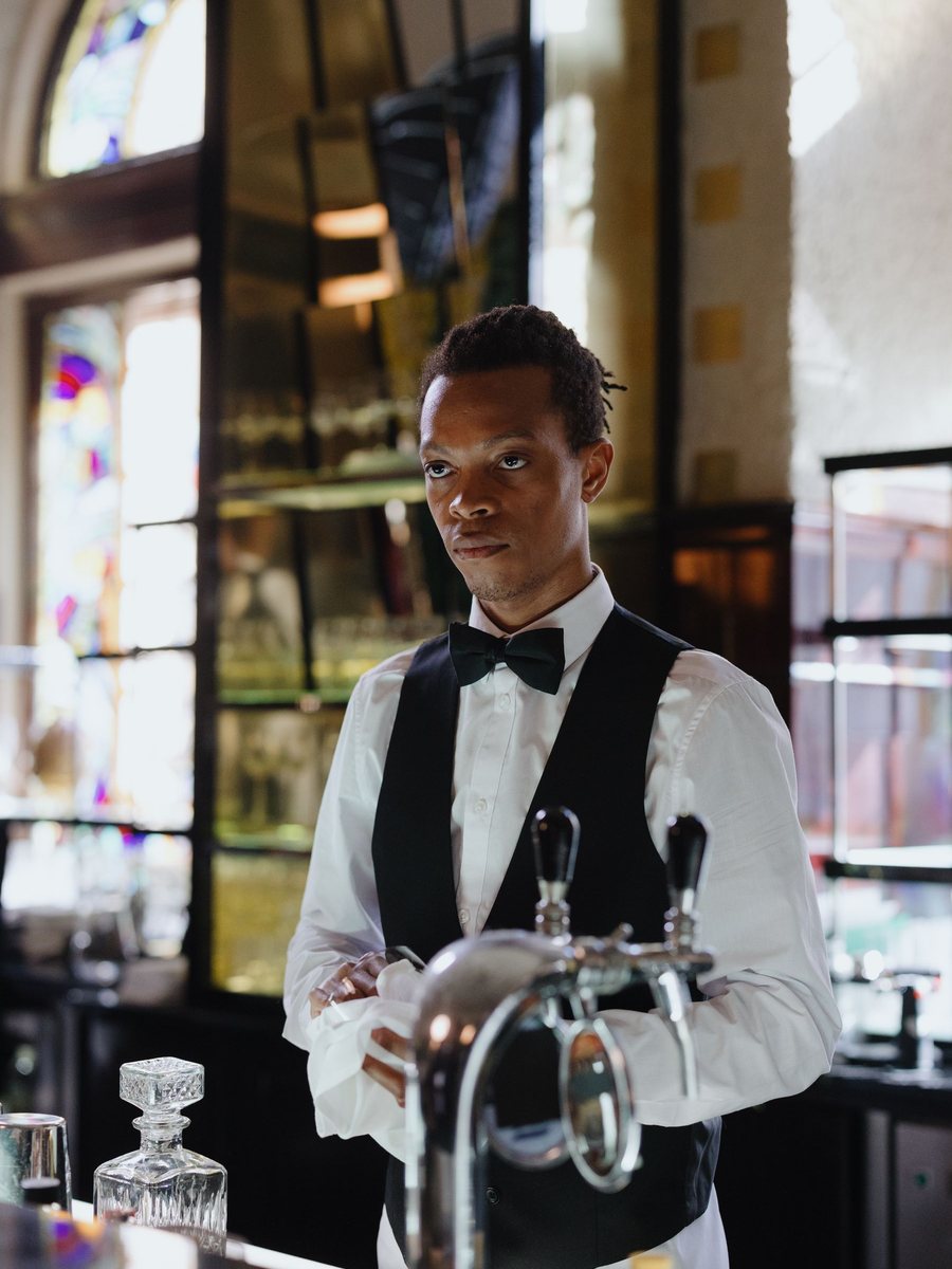 A bartender in a black vest and bow tie polishes a glass