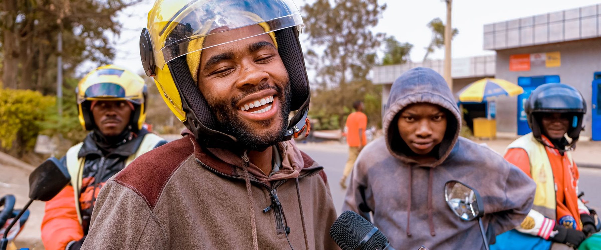 A smiling young man in a yellow helmet among other motorcycle riders at a boda boda stage