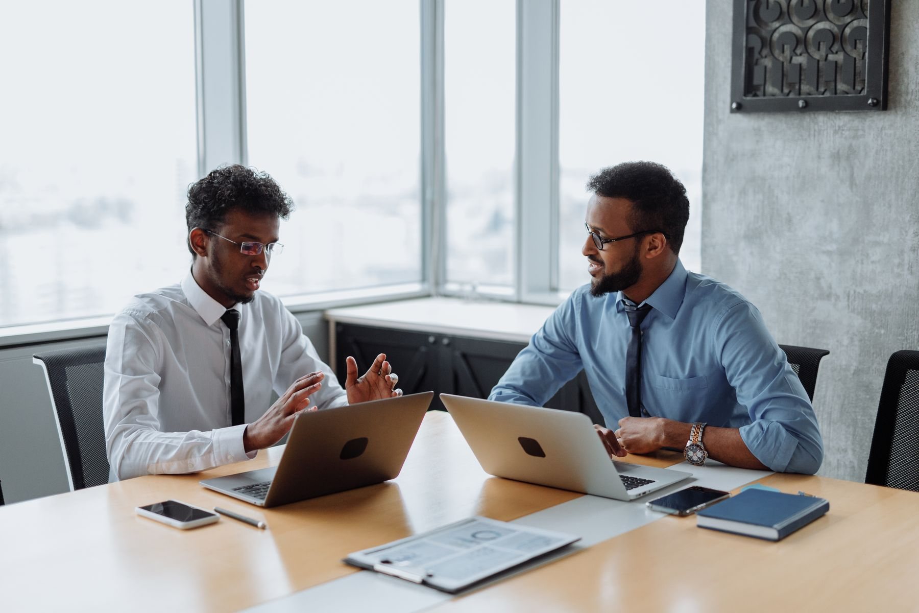 Two African men in shirts and ties talk across a meeting table with laptops open in a bright office with tall windows